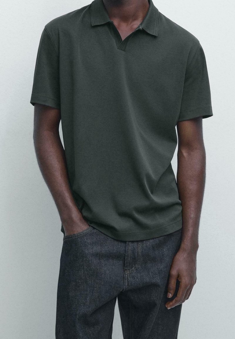Homme portant un polo vert foncé à manches courtes et un jean bleu foncé avec une main dans la poche, sur un fond uni clair.