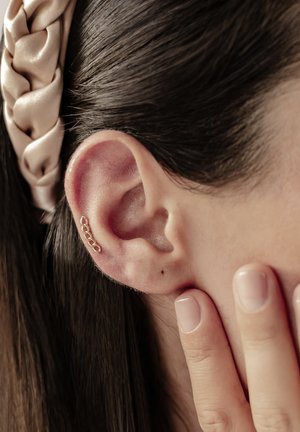 Gold ear cuff with a chain link design, attached to the outer ear. Hair is dark and glossy, with a fabric headband visible in background.
