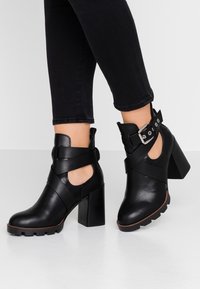 Botas de tobillo de cuero negro con un tacón grueso, que cuentan con lados recortados y una correa de hebilla ajustable. Textura suave y diseño audaz.