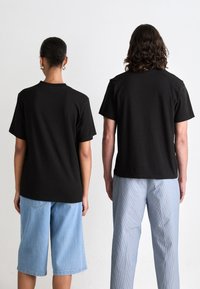 Sorte bomull T-skjorter båret av en person i blå denimshorts og en annen i stripete lyseblå bukser, sett fra bak.