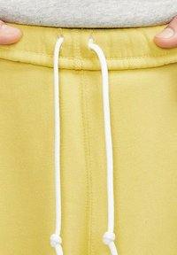 Gula sweatpants med en mjuk textur, som har en snörning med vita detaljer. Visar midjeband och tygdetaljer.