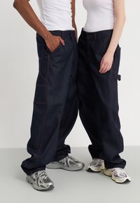 Pantalones cargo de mezclilla en azul oscuro con costuras rojas. Presenta múltiples bolsillos y un ajuste relajado. Combinados con zapatillas deportivas.