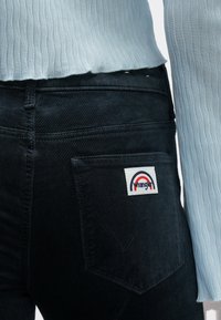 Calças de ganga pretas com acabamento texturizado, apresentando uma pequena etiqueta branca da Wrangler com um design de arco vermelho e azul no bolso traseiro.