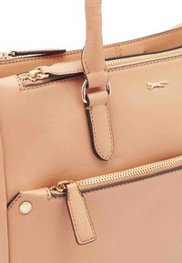 Tan leren handtas met goudkleurige hardware, voorzien van een ritsvak en gestikte accenten. Logo is op de zijkant gedrukt.