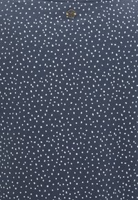 Mörkblått tyg med ett vitt prickmönster, med en metalllogotyp som accent vid bakhalslinjen, tillverkat av mjukt bomullsmaterial.