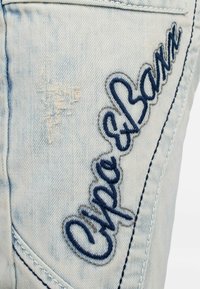 Jeans in denim azzurro chiaro con un motivo strappato e un logo ricamato in blu scuro, contornato da filo grigio sulla coscia.