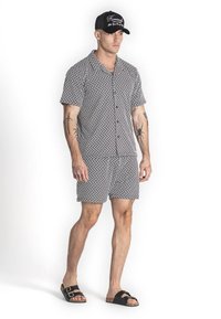 Camisa de manga corta con estampado geométrico en blanco y negro y pantalones cortos a juego, combinados con sandalias y una gorra negra con texto en blanco.