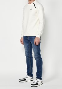 Felpa bianca con texture a waffle e spalla nera, abbinata a jeans slim-fit blu e sneakers bianco e nere.