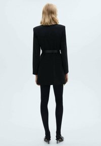 Schwarzes maßgeschneidertes Blazer-Kleid mit tailliertem Schnitt, langen Ärmeln und glatter Textur. Kombiniert mit schwarzen Strumpfhosen und Absätzen.