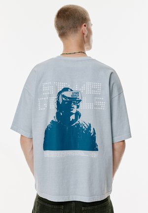 Młody mężczyzna z krótkimi włosami ubrany w jasnoszary T-shirt z niebieską grafiką osoby w okularach VR oraz napisem "STWD GOALS" na plecach.