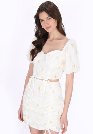 Młoda kobieta o długich brązowych włosach, ubrana w biały dwuczęściowy strój wyszywany żółtymi i białymi stokrotkami, na jednolitym tle.