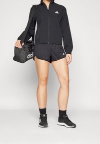 Sort træningsjakke med lynlås, hvide sidestropper; sorte shorts; sort fodtøj med hvide accenter; bærer en sort sportstaske.