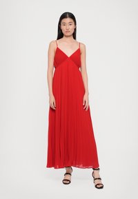 Červené plisované maxi šaty s tenkými ramienkami, majú V-výstrih a splývavú látku. Styling doplnený čiernymi sandálmi na platforme.