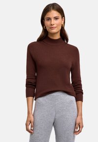 Eine Frau trägt einen braunen langen Pullover mit Rollkragen und hellgraue Hose. Sie steht mit entspannten Armen vor einem schlichten hellen Hintergrund.
