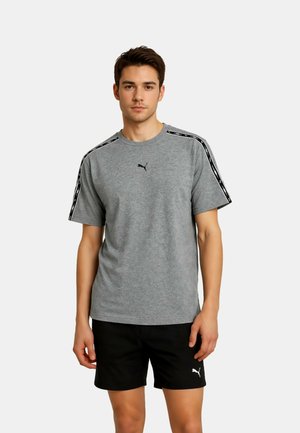 Młody mężczyzna w szarej koszulce Puma z czarnym logo i pasiastym detalem na ramieniu, w połączeniu z czarnymi szortami Puma, stojący na białym tle.