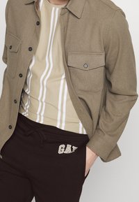 Braunes Wollmischgewebe-Hemd mit zwei Brusttaschen und schwarzen Knöpfen, über einem beige-weiß gestreiften T-Shirt getragen, ergänzt durch dunkle Sweatpants.