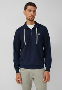 Sudadera de cuello redondo azul marino con media cremallera, cordones blancos y un pequeño parche con el logo en el pecho; puños y dobladillo acanalados.