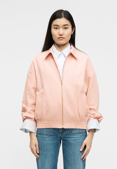 Νεαρό άτομο με μακριά μαύρα μαλλιά, φορώντας ένα ανοιχτό ροζ μπουφάν με φερμουάρ πάνω από μια ριγέ μπλούζα και μπλε τζιν, στέκεται μπροστά σε μια απλή φόντο.