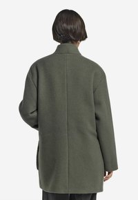 Olivgrön ullrock med krage, oversized passform, sänkta axlar och en ren bakseam, med sidofickor.