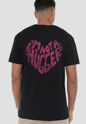 Mann trägt ein schwarzes T-Shirt mit pinkfarbenem Text in Herzform, der "Ich bin kein Umarmungstyp" lautet, von hinten gesehen.