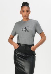 Pilka medvilnė marškinėliai su juodu "Calvin Klein" logotipu ir "ck" dizainu. Dėvima kartu su juoda odine sijonu, rankos į kišenes.