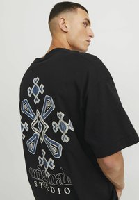 T-shirt nero a maniche corte con intricati ricami geometrici blu e bianchi sulla schiena con il testo "original STUDIO."