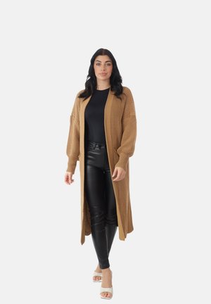 Donna in piedi che indossa un lungo cardigan beige in maglia, top nero, pantaloni in pelle neri e tacchi bianchi open toe su uno sfondo chiaro e semplice.