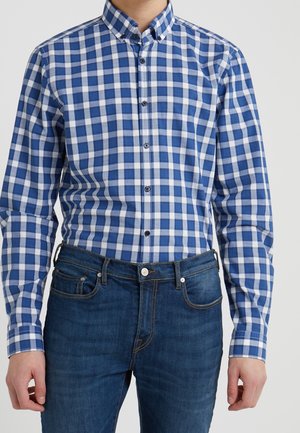 Homme portant une chemise à carreaux bleue et blanche boutonnée, rentrée dans un jean bleu foncé, sur un fond uni.