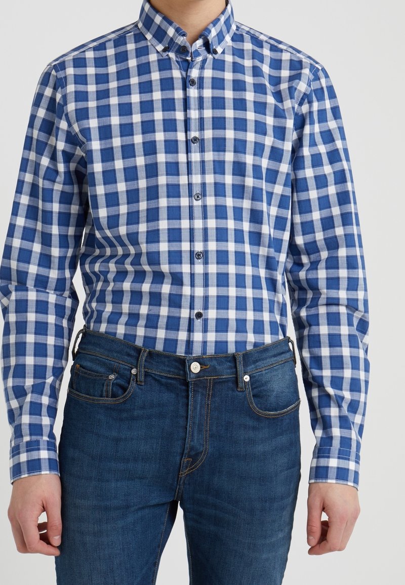 Man draagt een blauw-wit geruit overhemd ingestopt in donkerblauwe spijkerbroek tegen een effen achtergrond.