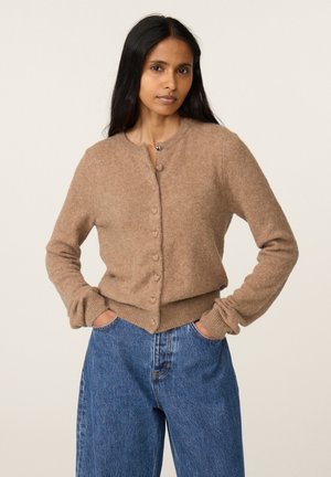 Frau mit langen schwarzen Haaren trägt eine hellbraune, geknöpfte Strickjacke und blaue Jeans, steht mit den Händen in den Taschen vor einem schlichten Hintergrund.