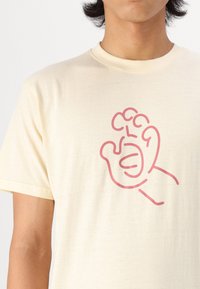 Crème t-shirt gemaakt van zachte stof met een grote, roze, omtrekillustratie van een hand met gestileerde vingers en een golvende element.