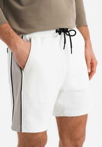 Hvide shorts med sorte side-striber. Lavet af blødt stof, med en elastisk talje og en sort snor samt sidelommer.