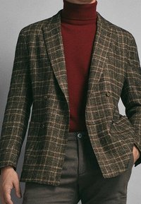 Blazer a scacchi marrone con chiusura doppiopetto e risvolto, indossato su un dolcevita color bordeaux; presenta un tessuto testurizzato e bottoni a vista.