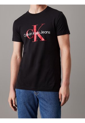Mężczyzna noszący czarną koszulkę Calvin Klein Jeans z dużym czerwono-białym logo i niebieskie jeansy na szarym tle.