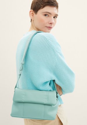 Mintgrüne Leder-Umhängetasche mit Klappdeckel, dezentem Steppdetail und verstellbarem Trageriemen, kombiniert mit einem hellblauen Strickpullover.