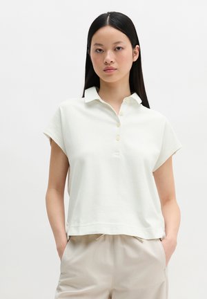 Fiatal nő hosszú fekete hajjal, laza fehér rövid ujjú galléros pólót és bézs nadrágot visel, kezét a zsebében tartva.