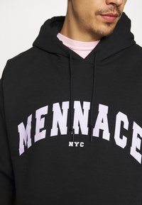 Homme portant un sweat à capuche noir avec texte blanc "MENNACE NYC", visible sous le menton et la moustache.