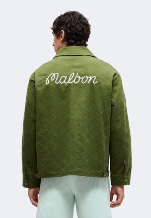 Mand iført grøn quiltet jakke med "Malbon" broderet hvidt på ryggen, kombineret med lyse shorts, set bagfra.