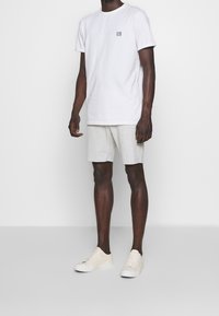 Man wearing white short-sleeve t-shirt with small logo, light beige shorts, and cream-colored sneakers against plain background.