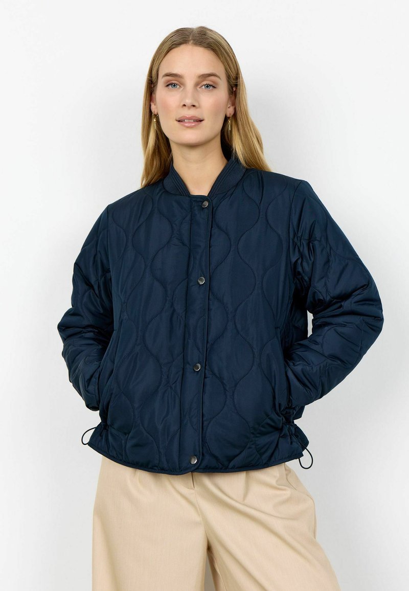 Žena s dlhými blond vlasmi v tmavomodrej prešívanej bunde so zapínaním na cvoky a béžových širokých nohaviciach, stojaca s rukami vreckách.