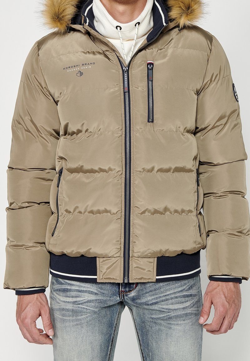 Doudoune beige avec capuche bordée de fourrure, poches zippées et poignets côtelés bleu marine, portée sur un sweat à capuche blanc avec un jean délavé clair.