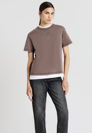 T-shirt marrone a maniche corte con finitura texture, sovrapposto a una camicia bianca, abbinato a pantaloni in denim grigio scuro. Design semplice con collo a girocollo.