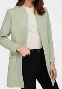 Abrigo de espiga verde claro con cuello mandarín, mangas largas y bolsillos laterales, superpuesto a un suéter crema y pantalones negros.