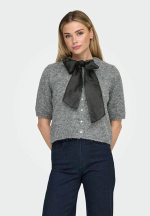 Femme portant un cardigan en maille à manches courtes gris avec des boutons en perle et un grand nœud noir au col, associé à un jean bleu foncé.