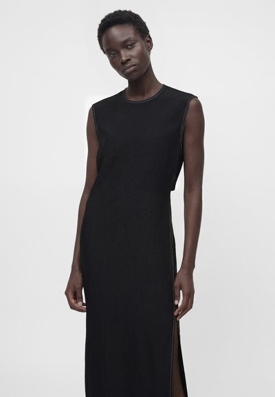 Γυναίκα που φοράει μαύρο αμάνικο φόρεμα με λευκές ραφές και πλαϊνή σκίσιμο, στέκεται μπροστά σε μονόχρωμο γκρι φόντο.