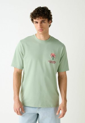 Fiatal férfi göndör hajjal, világoszöld pólóban, amelyen rózsaszín pálmafa és a "California" felirat látható, világoskék farmerrel párosítva.