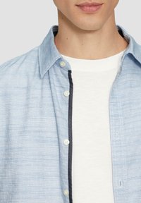 Niebieska w paski koszula z guzikami, z białą koszulką pod spodem. Koszula ma kontrastowy ciemny kołnierzyk i guziki.