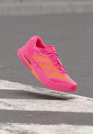 Zapatilla de running rosa brillante y naranja en el aire sobre un paso de peatones, con pequeños fragmentos de escombros dispersos alrededor.