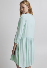 Rochie verde mentă cu mâneci până la trei sferturi, talie strânsă și fustă fluidă. Textură de material neted, design simplu, fără modele proeminente.