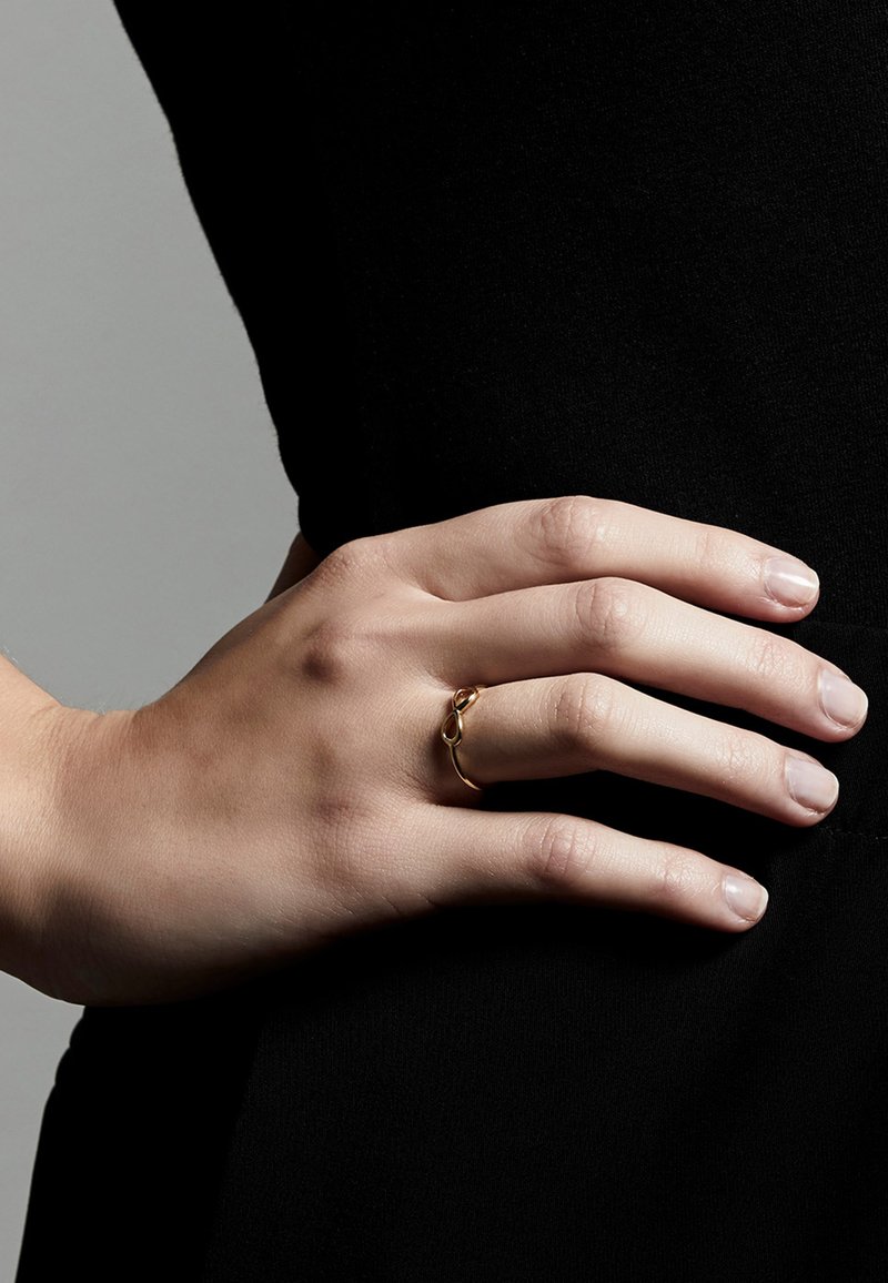 Goldring mit minimalistischem Design und einem sich überschneidenden Schleifendetail. Glatte Oberfläche, getragen an einer Hand vor einem dunklen Hintergrund.
